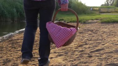 Kadının biri klasik piknik sepetiyle göl kıyısında yürüyor. 