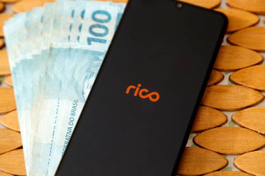Minas Gerais, Brezilya - 19 Nisan 2021: Rico broker marka akıllı telefon ekranı ve birkaç Brezilyalı gerçek banknot