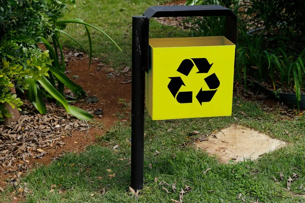 yellow metal trash can with recycling symbol in green public area and garden - garbage collection