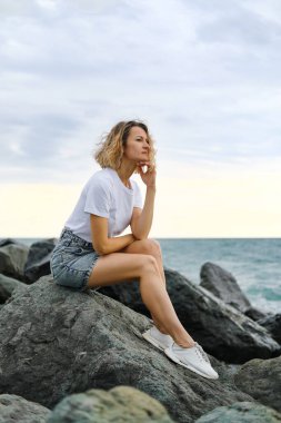 35 yaşında, genç, beyaz, sarışın bir kadın deniz kıyısındaki kayalarda oturuyor. Zümrüt renkli su, taşlara vuran dalgalar. Kadın meditasyon yapıyor ve rahatlıyor..