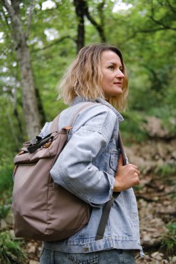 A young Caucasian woman walks along a forest mountain road. Casual blue denim clothes and backpack. Travel, healthy lifestyle and freedom.