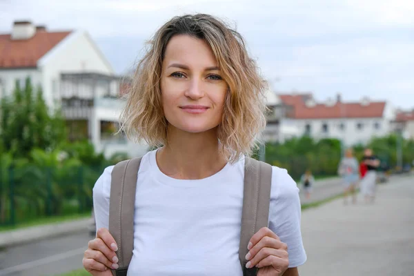 Mooie glimlachende krullende blanke vrouw in casual look loopt vol vertrouwen door de straat met rugzak. Wit t-short en blauw denim rokje. Levensvreugde, actieve levensstijl, modeblogger. — Stockfoto