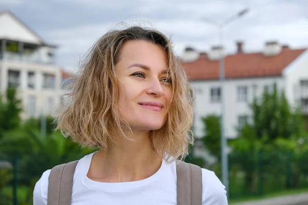 Mooie glimlachende krullende blanke vrouw in casual look loopt vol vertrouwen door de straat met rugzak. Wit t-short en blauw denim rokje. Levensvreugde, actieve levensstijl, modeblogger. — Stockfoto