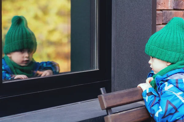 小さな子供が通りから窓の外を見ている 少年は鏡の窓に映る姿を見て — ストック写真