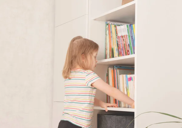 女の子は家で本棚の本を選ぶ. — ストック写真