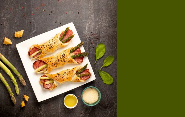 Baked green asparagus with ham and cheese in puff pastry sprinkled with sesame seeds and green leaves. Light gray stone background. top view. Free space for your text.