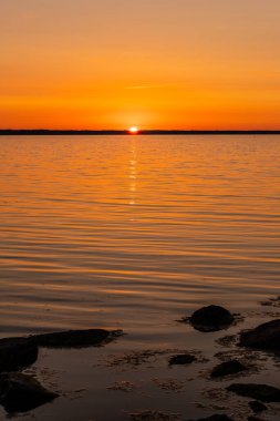 Altın güneş denizin üzerinde batıyor. Kayalar, deniz, güneş, ufukta orman silueti ve gökyüzünde beyaz kontrastlar bırakan jetlerle turuncu günbatımına dikey görüş. Gün batımı suyu ve gökyüzünü turuncu yapar.