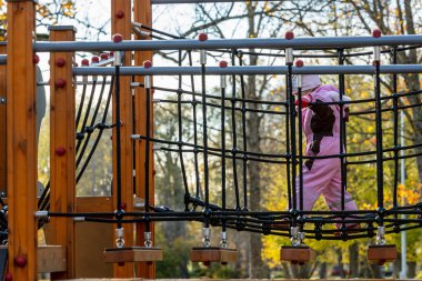 Pembe kışlık giyinmiş küçük bir kız, pembe şapkalı ve kırmızı eldivenli güneşli sonbahar gününde parktaki köprüde yürüyor. Çocuk parkında ağlarla oynayan genç bir kız. Çocuk sallantılı asma halat köprüsünde yürüyor.