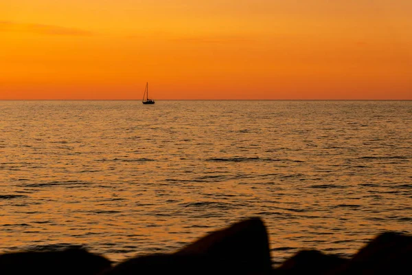 Denizde turuncu ve sarı günbatımı ve sol tarafında yatın silueti var. Alacakaranlıkta denizde yüzen bir teknenin silüeti. Costal rocks, yelkenli, günbatımı, deniz, altın saat, siluetler. 