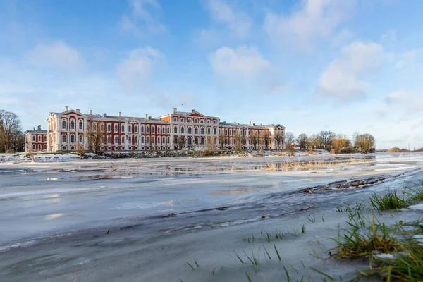 Güneşli kış gününde, donmuş nehir kıyısındaki Barok malikanesinin alçak açılı görüntüsü. Jelgava Sarayı soğuk kış gününde nehirde.