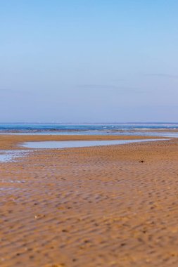 Kuzeydeki koyu sarı kumlu sahil şeridinin dikey fotoğrafı kısmen suyla kaplı, uzak deniz, kasaba ve açık mavi gökyüzü. Bahar başında Letonya, Jurmala 'daki Majori plajında boş bir tatil yeri.. 