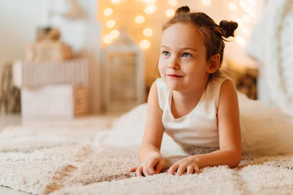 Ein lustiges kleines Mädchen in einem weißen Kleid liegt auf einer Matte im Schlafzimmer — Stockfoto