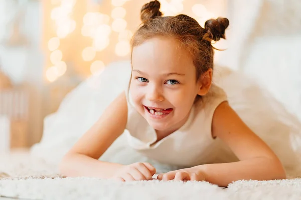 Uma menina bonita em um vestido branco encontra-se em um tapete no quarto — Fotografia de Stock