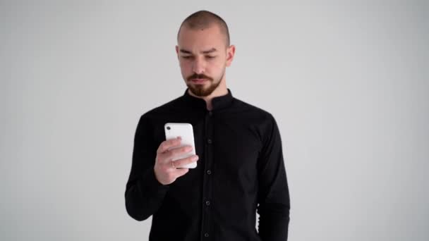 若いハンサムな髭を生やした男が黒いシャツに白い背景に携帯電話を手に持っていて何が起こっているのか理解していない — ストック動画