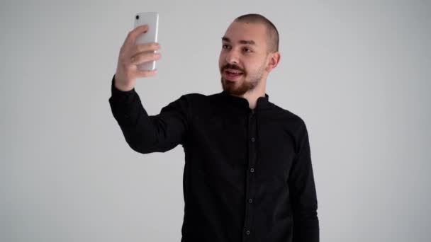 黒のシャツを着た若いハンサムな髭の男が白い背景に携帯電話を手にビデオ通話をして — ストック動画