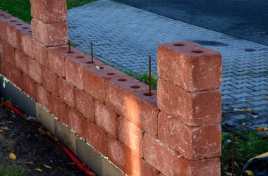 Beton döşemeden çit yapımı. Kırmızı tuğlaların içinde çelik telleri yerleştirmek ve beton çamurla doldurmak için delikler var. Görünür renklerden yapılmış katı ve ucuz çitler. 