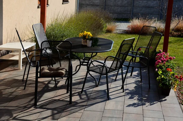 the terrace at the house in a beautiful garden is equipped with a garden furniture made wire metal chairs and an oval table. gray concrete brick terrace. flowering and ornamental grasses, flower pot
