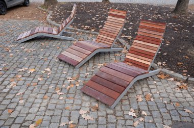 Meydandaki ağaçların altındaki kaldırımda sallanan sandalye. Parktaki tek bir kişi için tropikal ahşaptan yapılmış ahşap sandalyeler. Kahverengi kalaslar, çatallar ve çelik çerçevelerden yapılmışlar.