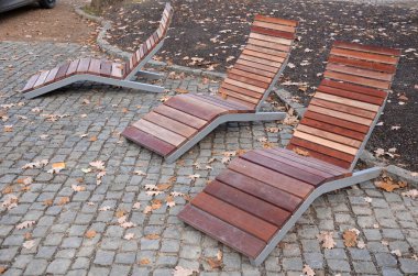 Meydandaki ağaçların altındaki kaldırımda sallanan sandalye. Parktaki tek bir kişi için tropikal ahşaptan yapılmış ahşap sandalyeler. Kahverengi kalaslar, çatallar ve çelik çerçevelerden yapılmışlar.