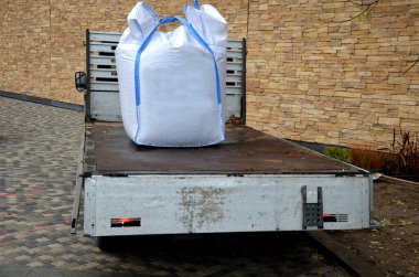 Daha küçük bir kamyona büyük beyaz plastik torbalar dolusu çatı döşemesi yüklüyorlar. Vinç havada, kancanın saplarının yanında bir metre küp ağırlığında çakıl tutuyor..