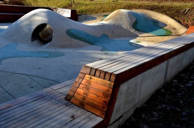 Tepeli, mavi-mavi-yeşil kauçuk manzara ve sahada renkli noktalar. Tepenin içinden paslanmaz çelik tünel. ahşap kaplanmış eğimli köşe bank istinat duvarı