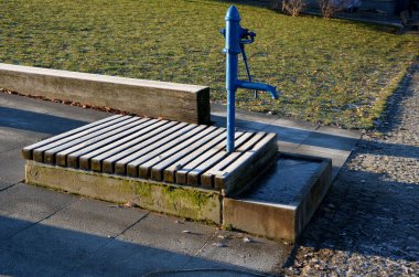 Mavi metal retro, parlak ahşap kapaklı. Su el pompasından taş yalağa akar. Bahçe tezgahı şekilli prizma çimenlik park mimarisi, dayanıklılık, ucuz