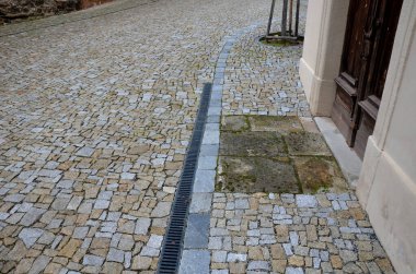 Kabaca düzensiz asfalt istiflenmiş yuvarlak ağaç kaseleri. Tarihi bir binanın kahverengi duvarları ve sokaktan uzaklaşan boylamasına bir kanal. Yeni dikilmiş bir ağacın drenaj borusu.