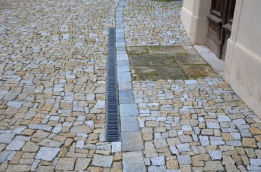 Kabaca düzensiz asfalt istiflenmiş yuvarlak ağaç kaseleri. Tarihi bir binanın kahverengi duvarları ve sokaktan uzaklaşan boylamasına bir kanal. Yeni dikilmiş bir ağacın drenaj borusu.