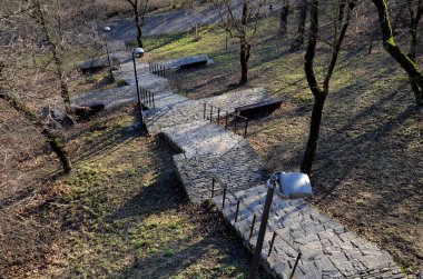 Dinlenme yerleri olan merdivenler. Parkta yamaçtan doğruca gölete gidiyor. Siyah metal korkulukları tutuyor. Taş doğa, beton yollar ve merdivenler. Lambalar ve ahşap banklar