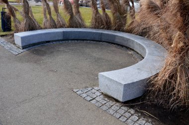 Japon uzun kuru otları kış için demetlere bağlandı. Parktaki granit bankın etrafını saran çiçek tarhının kemerinde. Bank at nalı gibi kıvrılmış, kaldırımın kenarında pürüzsüz bir yüzeyi var.