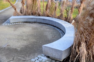 Japon uzun kuru otları kış için demetlere bağlandı. Parktaki granit bankın etrafını saran çiçek tarhının kemerinde. Bank at nalı gibi kıvrılmış, kaldırımın kenarında pürüzsüz bir yüzeyi var.