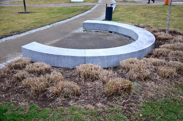 Japon uzun kuru otları kış için demetlere bağlandı. Parktaki granit bankın etrafını saran çiçek tarhının kemerinde. Bank at nalı gibi kıvrılmış, kaldırımın kenarında pürüzsüz bir yüzeyi var.