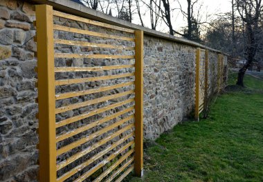 Taş duvarın yanındaki çimenlikte tek başına duran bitkilere tırmanmak için saraylar. Sürünen gül. Sarı renkten yatay ahşap bir duvar. Bahçedeki kare merdiven. Tırmanma bitkileri için dizayn.