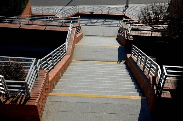 stairs and railings of exposed brick smooth surface. Metal railings and benches in the corners of the U-shaped brick retaining walls. The recessed lights illuminate the pedestrians\' feet under  feet