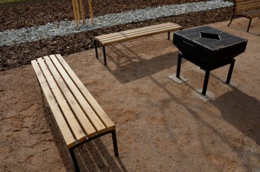 Pergola, bahçe köşkünde metal bir ızgara var. Parktaki umumi barbekü yerinde. Ahşap kamp masaları. Parktaki bej çakıl tarlalarının etrafındaki çimenler, yeni, bahar, siyah