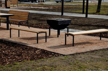 Pergola, bahçe köşkünde metal bir ızgara var. Parktaki umumi barbekü yerinde. Ahşap kamp masaları. Parktaki bej çakıl tarlalarının etrafındaki çimenler, yeni, bahar, siyah