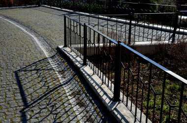 Asfaltlı kaldırım oluğu ve gri metal süslemeli korkuluklar. Lambanın gölgesi ve siyah direği şehrin sokaklarında eski gaz lambalarının yanında.