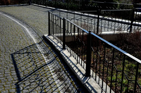 Asfaltlı kaldırım oluğu ve gri metal süslemeli korkuluklar. Lambanın gölgesi ve siyah direği şehrin sokaklarında eski gaz lambalarının yanında.