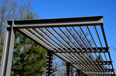 Otobüs durağının metal inşaatı, çardak pergola barınağı. Çatı bitkilere tırmanmak için tasarlandı. L-boyalı kirişler ve L şeklinde kaburgalar park, gökyüzü, mavi çizgili panjurlar.