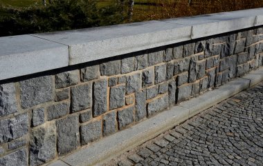 Geniş taş levhalardan yapılmış granit parmaklıklar ve gri bloklardan yapılmış taş duvarlar. Gezinti güvertesinde oturma duvarı görevi görüyor. Arka planda çalılar büyür.