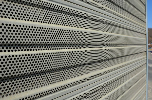 noise barrier made of metal perforated sheet metal slats. gray and silver protective fencing separates housing from a busy noisy road. the porous material shatters tire noise well.