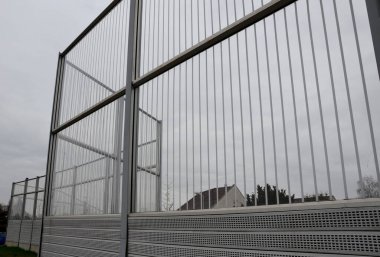 Metal delikli metal levhalardan oluşan çizgili cam ses bariyeri. Gri ve gümüşi koruyucu çitler gürültülü bir yoldan evleri ayırıyor. Gözenekli madde lastik sesini iyi parçalar..