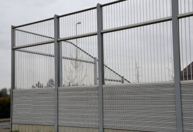 Metal delikli metal levhalardan oluşan çizgili cam ses bariyeri. Gri ve gümüşi koruyucu çitler gürültülü bir yoldan evleri ayırıyor. Gözenekli madde lastik sesini iyi parçalar..
