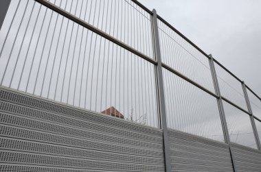 Metal delikli metal levhalardan oluşan çizgili cam ses bariyeri. Gri ve gümüşi koruyucu çitler gürültülü bir yoldan evleri ayırıyor. Gözenekli madde lastik sesini iyi parçalar..