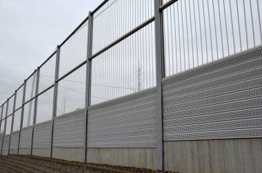 Metal delikli metal levhalardan oluşan çizgili cam ses bariyeri. Gri ve gümüşi koruyucu çitler gürültülü bir yoldan evleri ayırıyor. Gözenekli madde lastik sesini iyi parçalar..