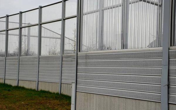striped glass noise barrier made of metal perforated sheet metal slats. gray and silver protective fencing separates housing from a busy noisy road. the porous material shatters tire noise well.
