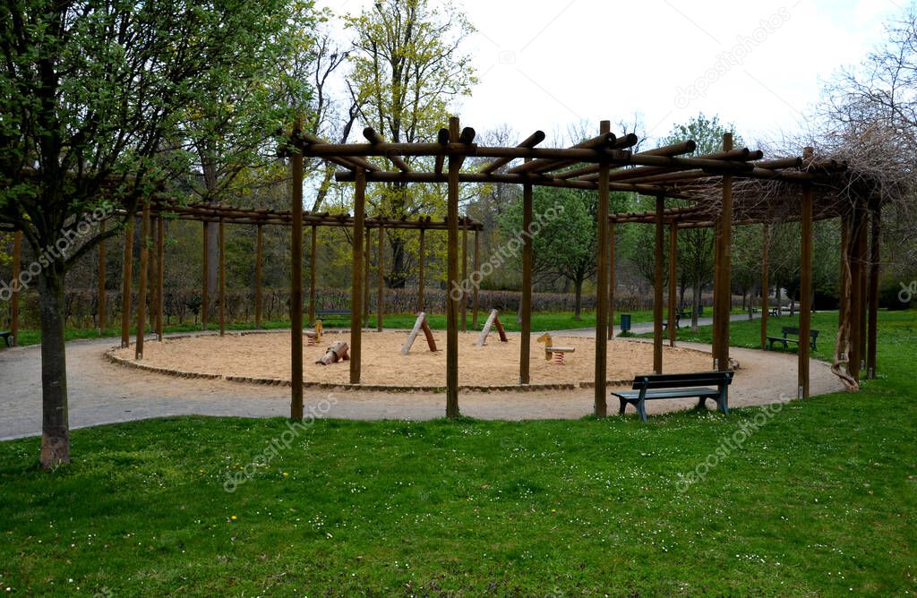 round pergola, trellises made of wooden poles around the sandpit with play elements for children. around is a path and lawns. benches for moms to rest. stakes in Japanese Chinese style