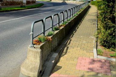 Meydanın yanındaki süs saksıları. Süslü çiçekli otlar. Tahta malzemeden yapılma saksı şeklindeki saksı. Kamp tezgahı, masa, beton, korkuluk, direk