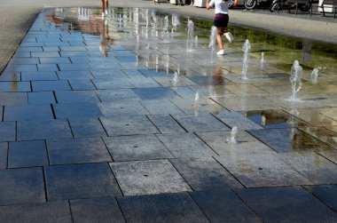 Meydanda, programa göre yukarı doğru püsküren düzenli bir ızgarada sprey hortumlar var. Kızlar şortlarıyla oynaşıp fıskiyede çıplak ayaklarıyla koşuyorlar.