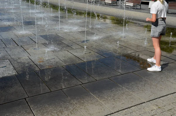 on the square there are spray nozzles in a regular grid which spray upwards according to the program. the girls are fooling around in their shorts and running naked legs through the fountain